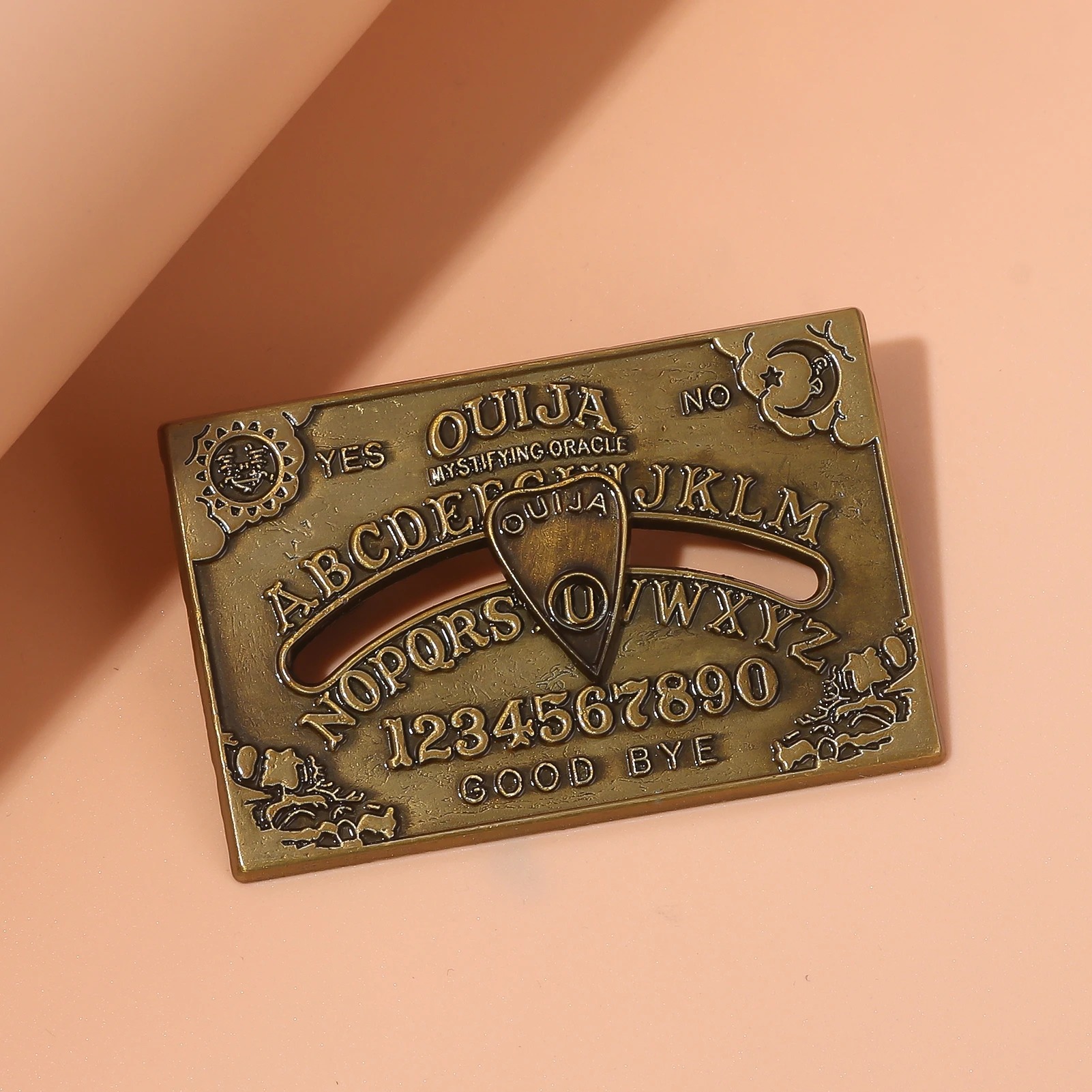 Broche do tabuleiro Ouija em bronze sobre fundo laranja e rosa, com sol e lua nos cantos