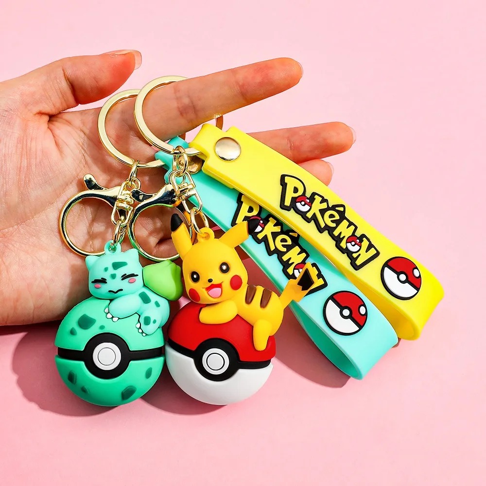 Mão segurando chaveiros do Bulbasaur e do Pikachu sobre Pokébolas, com alças coloridas