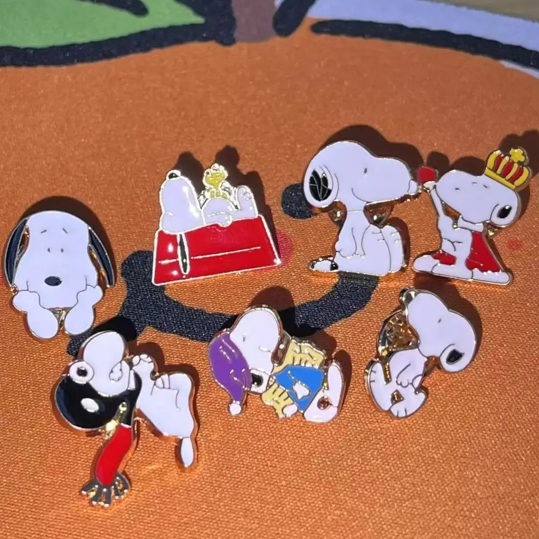 Kit de sete broches esmaltados do Snoopy, em poses diferentes, sobre fundo laranja