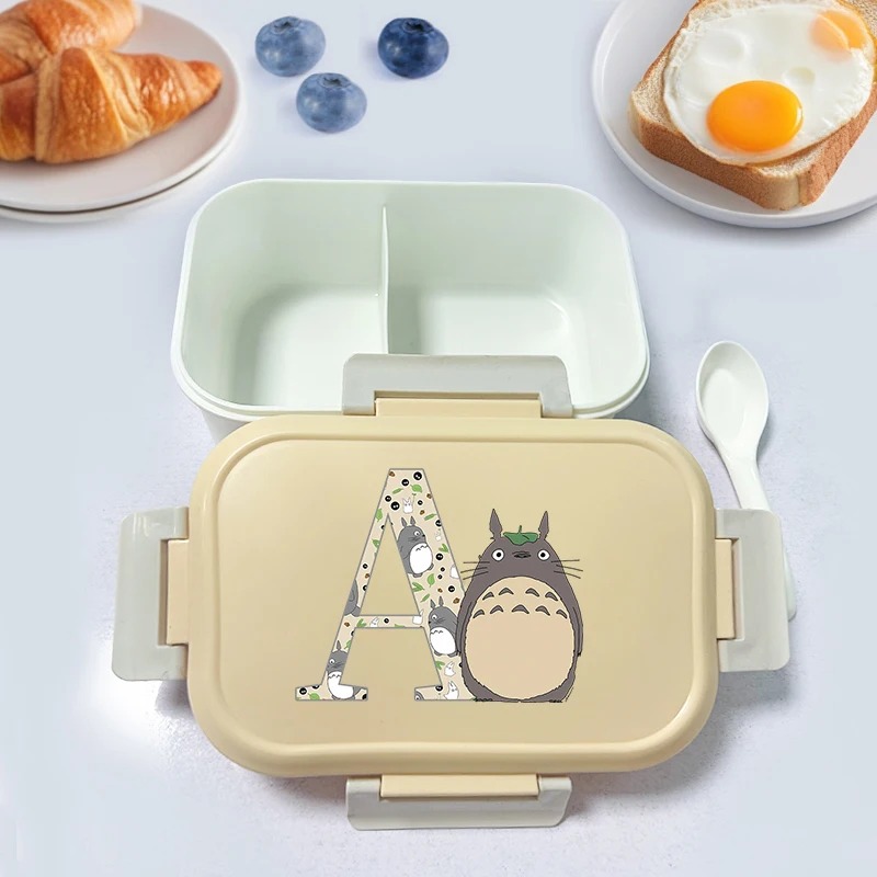 Marmita bento bege com o Totoro e a letra A estampada na tampa, ao lado de pão e ovo