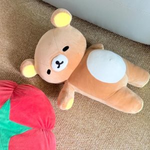 Pelúcia do Rilakkuma deitada sobre um tapete, urso marrom com barriga clara e orelhas amarelas